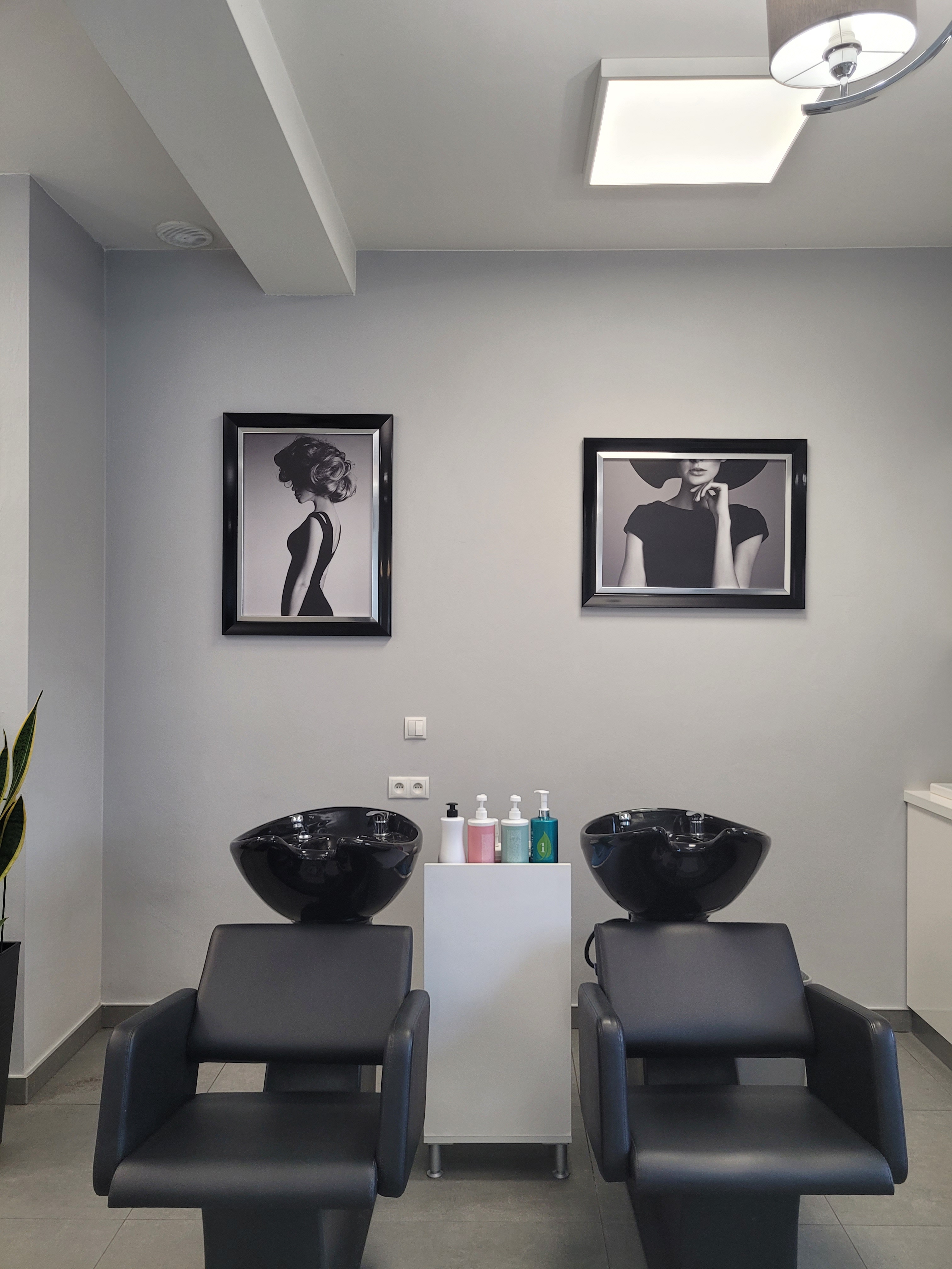 Czarne myjki fryzjerskie, szara ściana z czarno-białymi obrazami eleganckich pań, biała półka na szampony i odżywki.