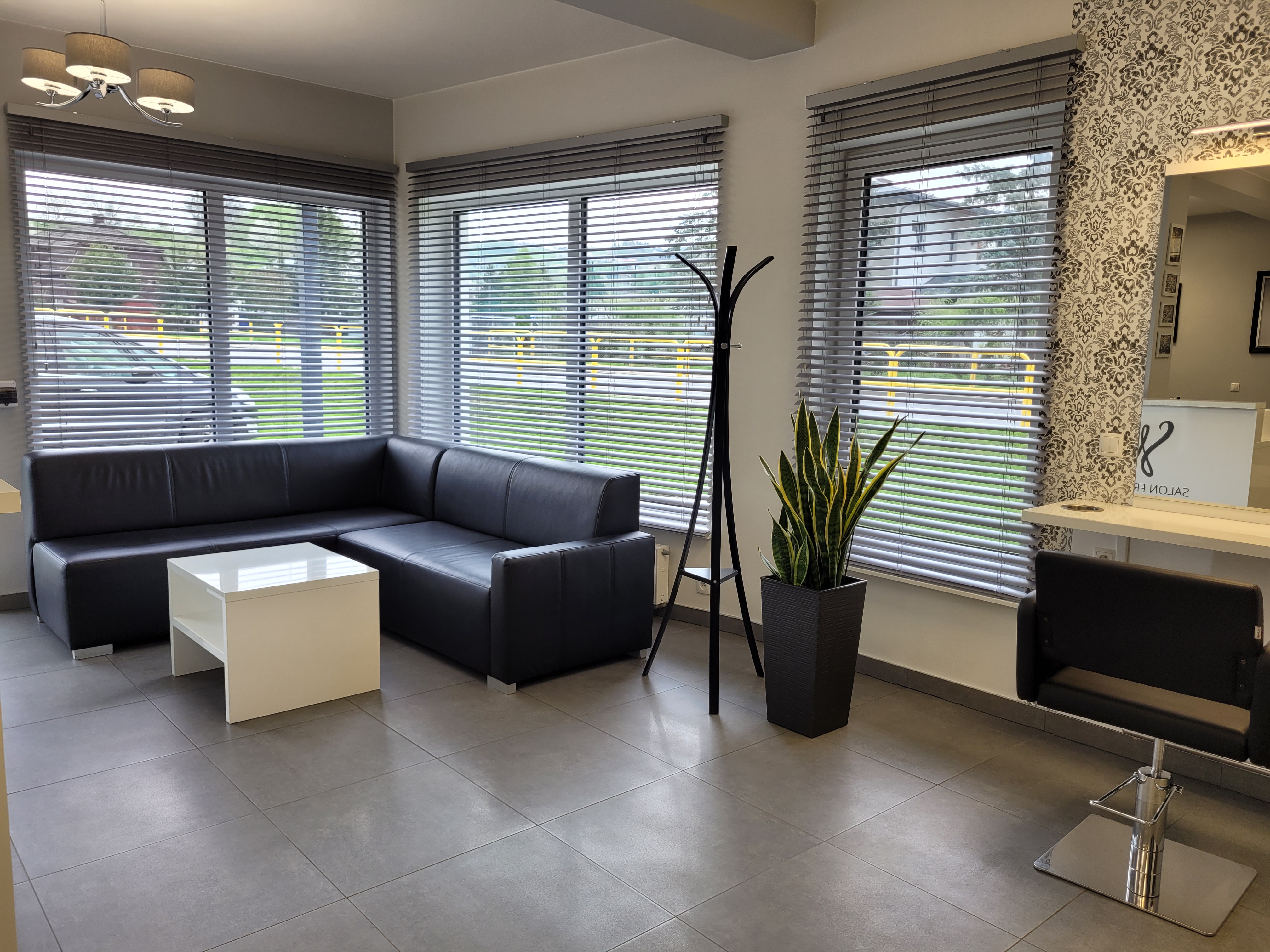 Aranżacja poczekalni w salonie fryzjerskim – projekt wnętrza z czarną kanapą, białym stolikiem i roletami, Nowy Sącz.